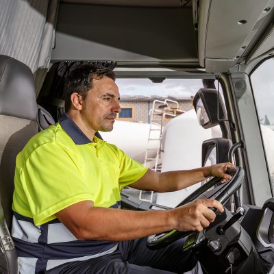 Side view of concentrated middle age male driver in lemon t shirt and curly hair sitting in tanker truck while driving in company compound during work hour