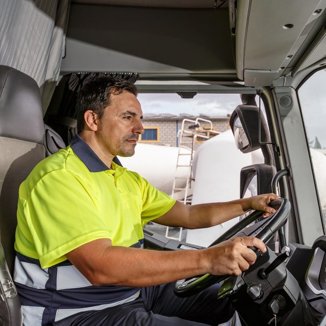Side view of concentrated middle age male driver in lemon t shirt and curly hair sitting in tanker truck while driving in company compound during work hour