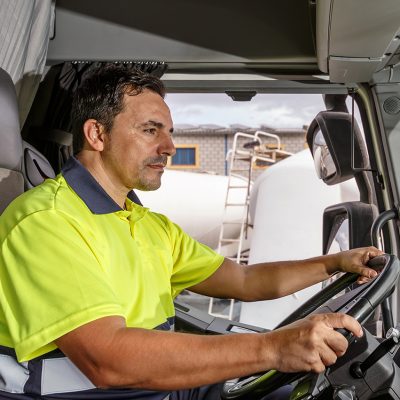 Side view of concentrated middle age male driver in lemon t shirt and curly hair sitting in tanker truck while driving in company compound during work hour